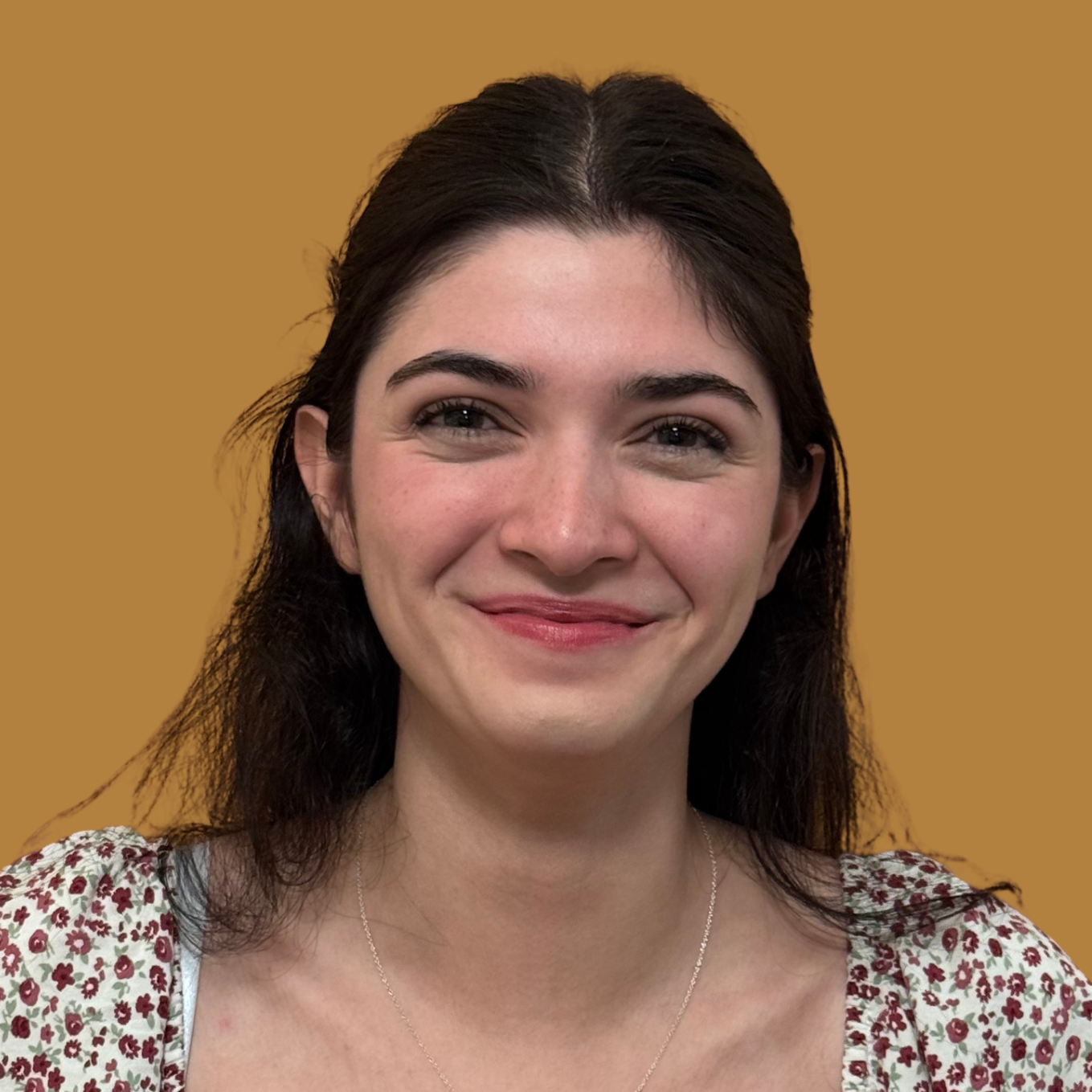 A woman with brown hair wearing a floral blouse and necklace stands in front of a yellow background.