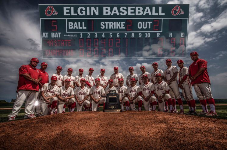 A baseball team poses for a group photo on the field in front of a scoreboard displaying the game status, with players wearing white uniforms and red caps.