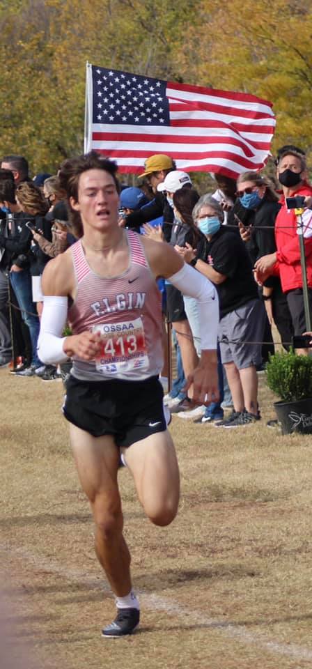 A male runner in athletic gear sprints towards the finish line, with a crowd cheering in the background and an American flag waving prominently.