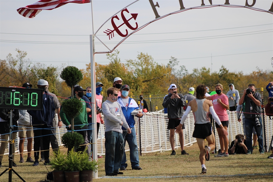 A runner crosses the finish line at a race, with a digital timer displaying 16:10 and spectators cheering in the background under a banner that reads 'Finish'.