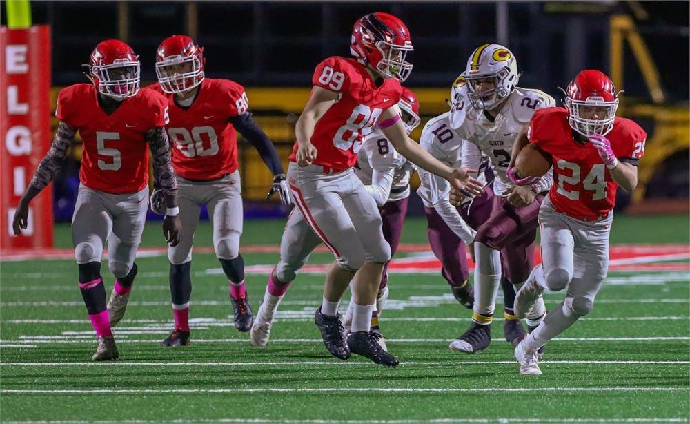 A group of football players in red uniforms are running on a field, with one player carrying the ball while being pursued by opponents in white and maroon uniforms.