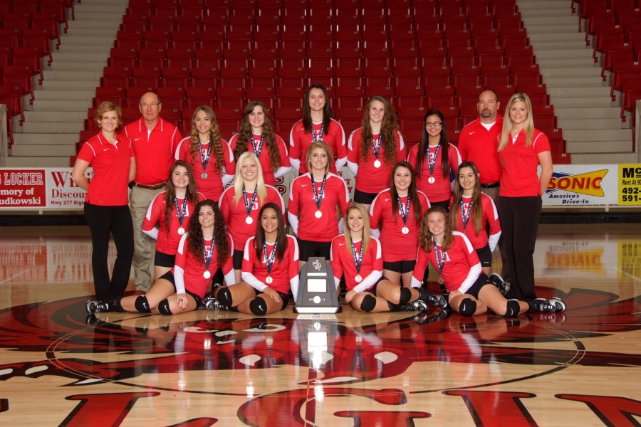 A volleyball team poses for a group photo on a gymnasium court, wearing red uniforms and holding a trophy, with coaches standing behind them.