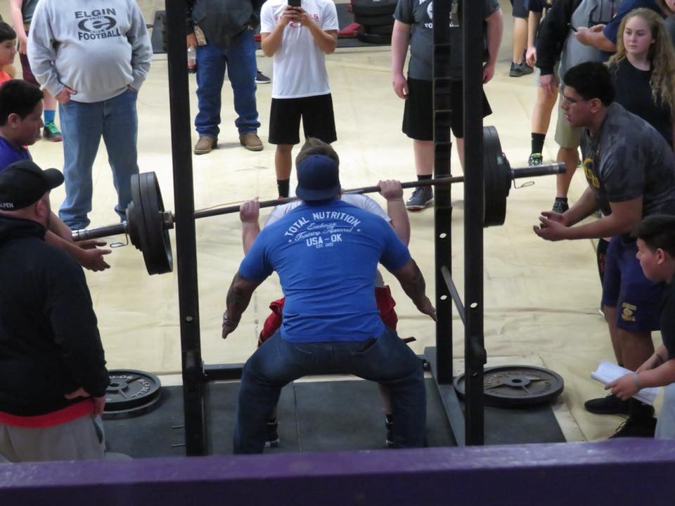 A weightlifter is performing a squat under the supervision of several spectators and coaches in a gym setting.