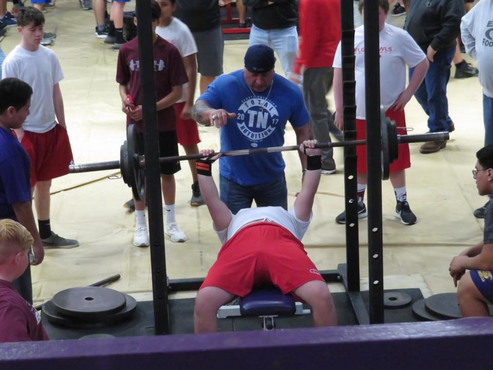 A group of people watches as a young man performs a bench press in a gym setting, with a trainer assisting him.