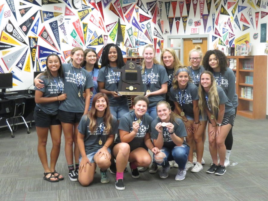 A group of young women in matching gray t-shirts pose together in a library, holding a trophy and celebrating their achievement, with colorful banners in the background.
