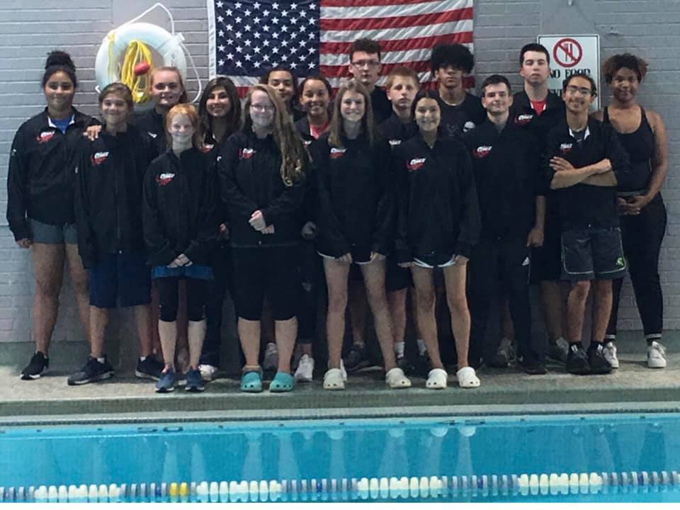 A group of swimmers in matching jackets poses together in front of an American flag and a swimming pool.