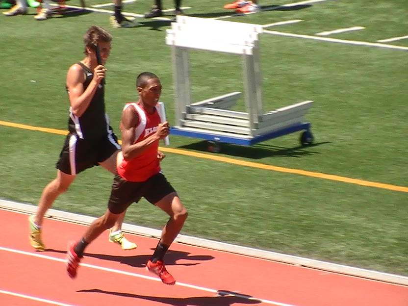 Two young male athletes are competing in a sprint race on a track, with one runner in a red uniform leading and another in a black uniform closely behind.