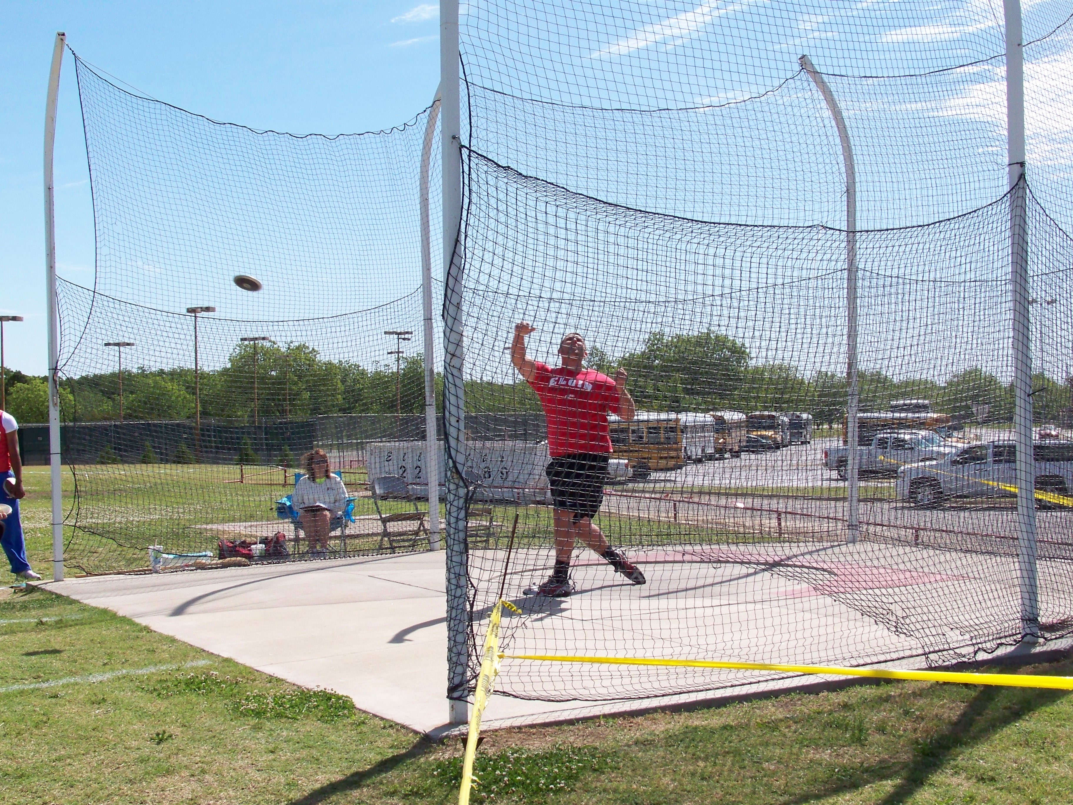 An athlete in a red shirt is in the process of throwing a discus within a fenced area, with spectators seated nearby and a clear blue sky above.