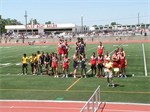 A group of athletes in colorful uniforms stands together on a track field, with spectators in the background and a stadium visible in the distance.