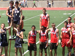 A group of male athletes stands on a podium celebrating their achievements at a track and field event, with some wearing medals and team uniforms in red and blue.