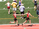 Two female athletes in red uniforms are passing a baton during a relay race on a track, with spectators in the background.