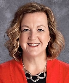 A smiling woman with curly hair wearing a red blazer and a necklace, set against a gray background.