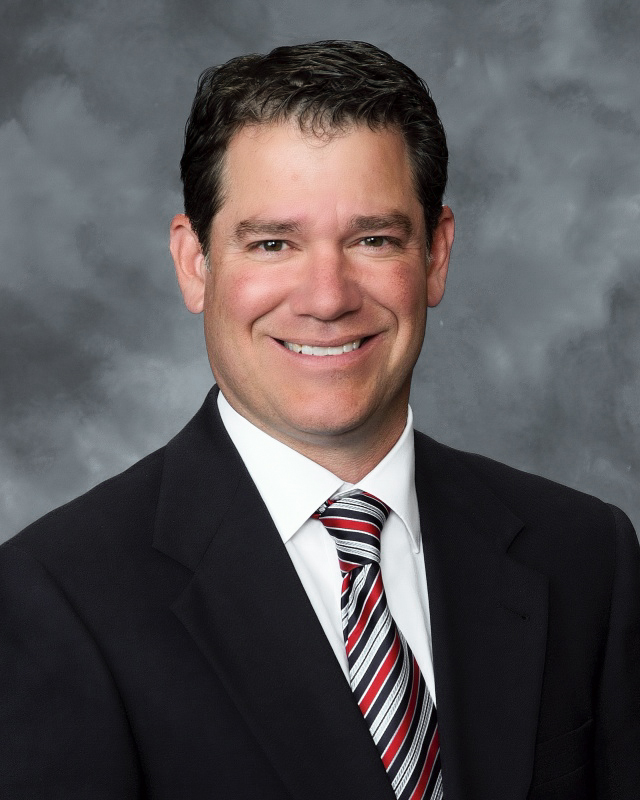 A smiling man in a black suit and striped tie poses against a gray background.