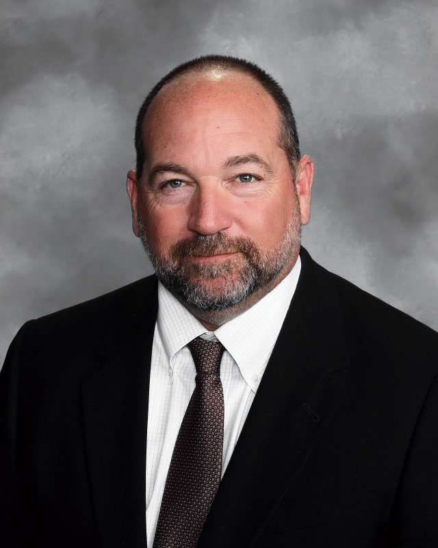 A man with a beard and short hair, wearing a black suit and a white shirt with a tie, poses against a gray background.