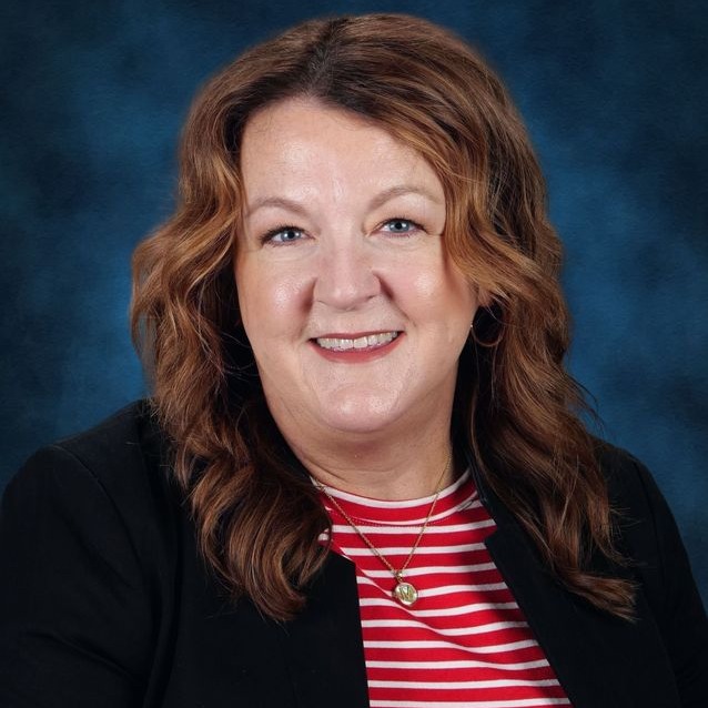 A smiling woman with curly hair wearing a red blazer and a necklace, set against a gray background.