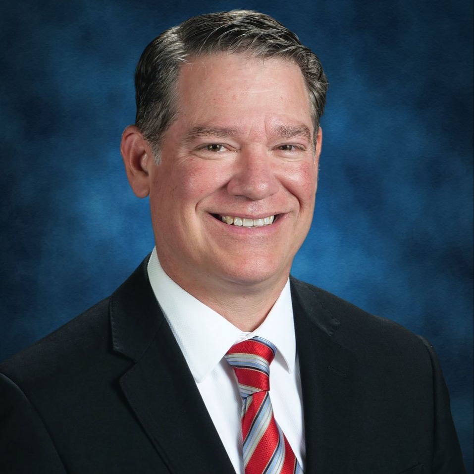 A smiling man in a black suit and striped tie poses against a gray background.