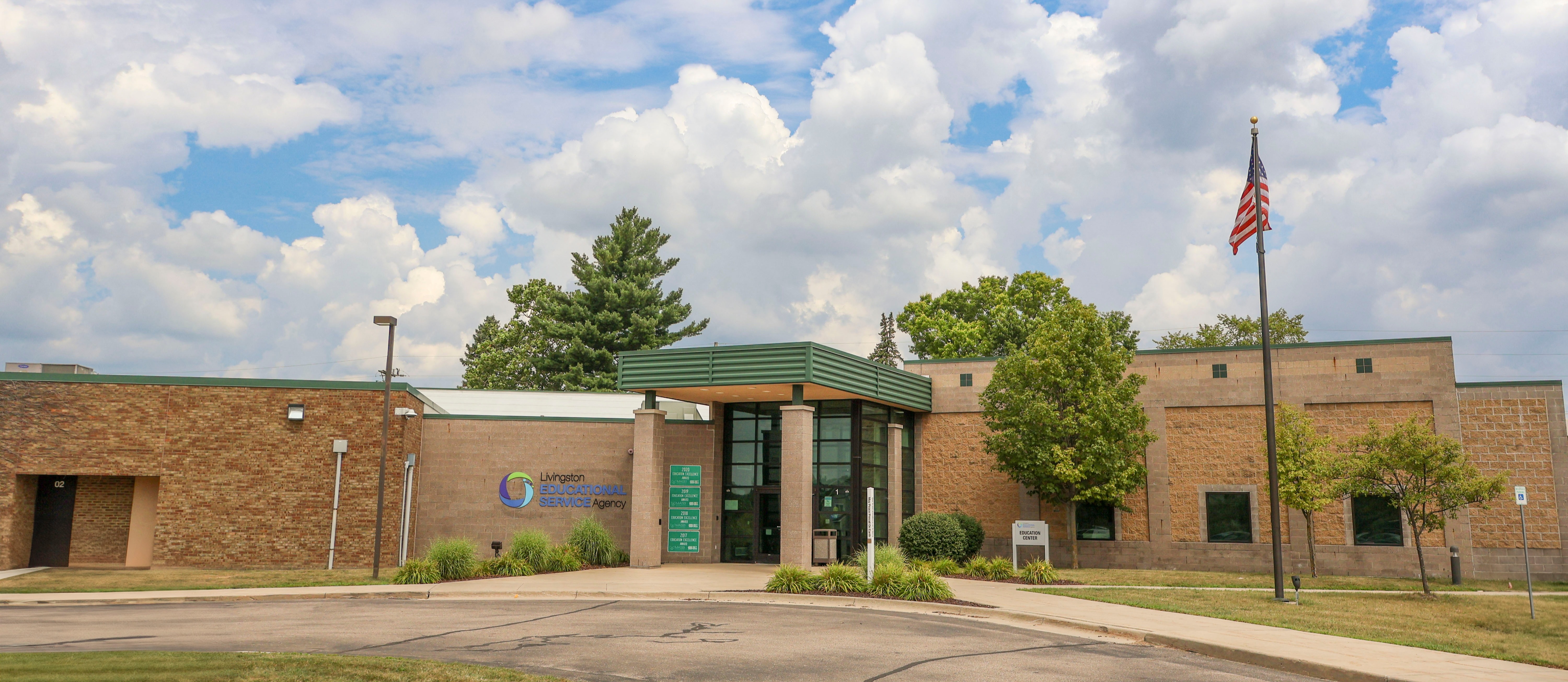 The exterior front entrance of the Livingston Educational Service Agency building featuring an American flag on a flagpole under a cloudy sky.