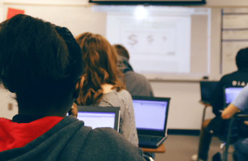 A classroom scene showing students seated at desks with laptops, facing a whiteboard displaying a mathematical equation.