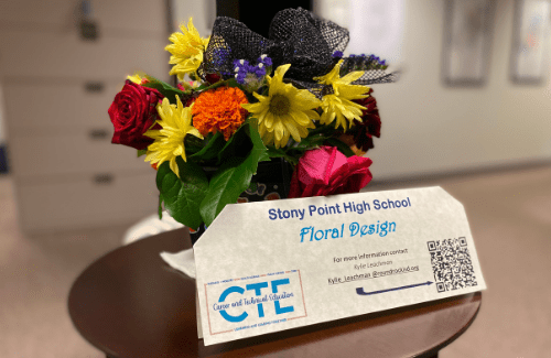 A colorful floral arrangement featuring various flowers, including roses and sunflowers, displayed on a table with a sign that reads 'Stony Point High School Floral Design' and includes a QR code.