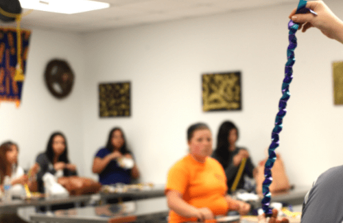 A person holds up a blue object in a classroom setting, while several individuals in the background are seated at tables, some engaged in conversation and others focused on their tasks.