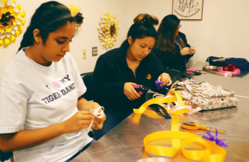 Two young women are engaged in a crafting activity, using scissors and colorful ribbons at a table, while another person is visible in the background.