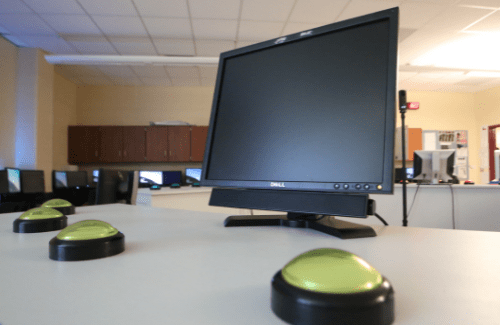 A computer monitor is positioned on a desk with several green buttons in the foreground, set in a classroom or office environment.