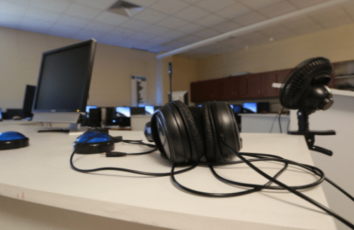 A pair of black headphones with tangled wires rests on a white table, with a computer monitor and other equipment visible in the background.