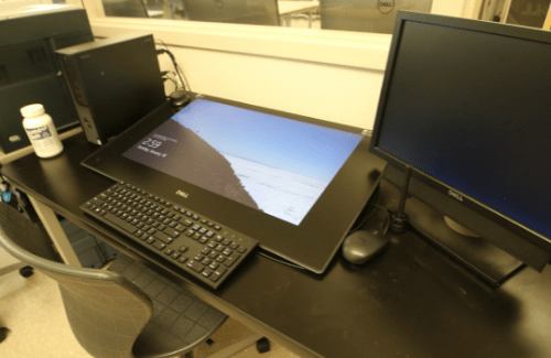 A workspace featuring a large touchscreen monitor, a standard computer monitor, a keyboard, and a mouse, all set on a black desk with a chair nearby.