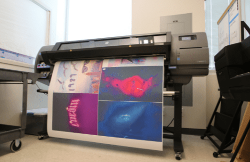 A large-format printer is shown in a room, with a partially printed sheet displaying colorful images emerging from the front.