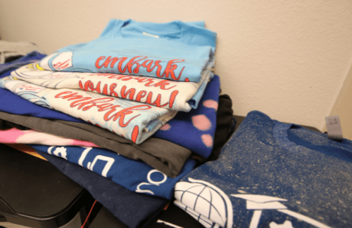 A stack of folded t-shirts in various colors and designs, with some featuring text and graphics, placed on a flat surface.