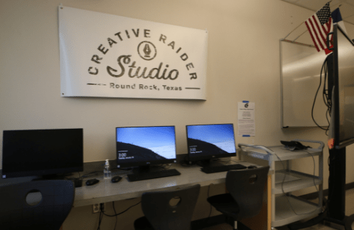 A workspace featuring three computers on a table, with a sign above that reads 'Creative Raider Studio' and an American flag in the background.