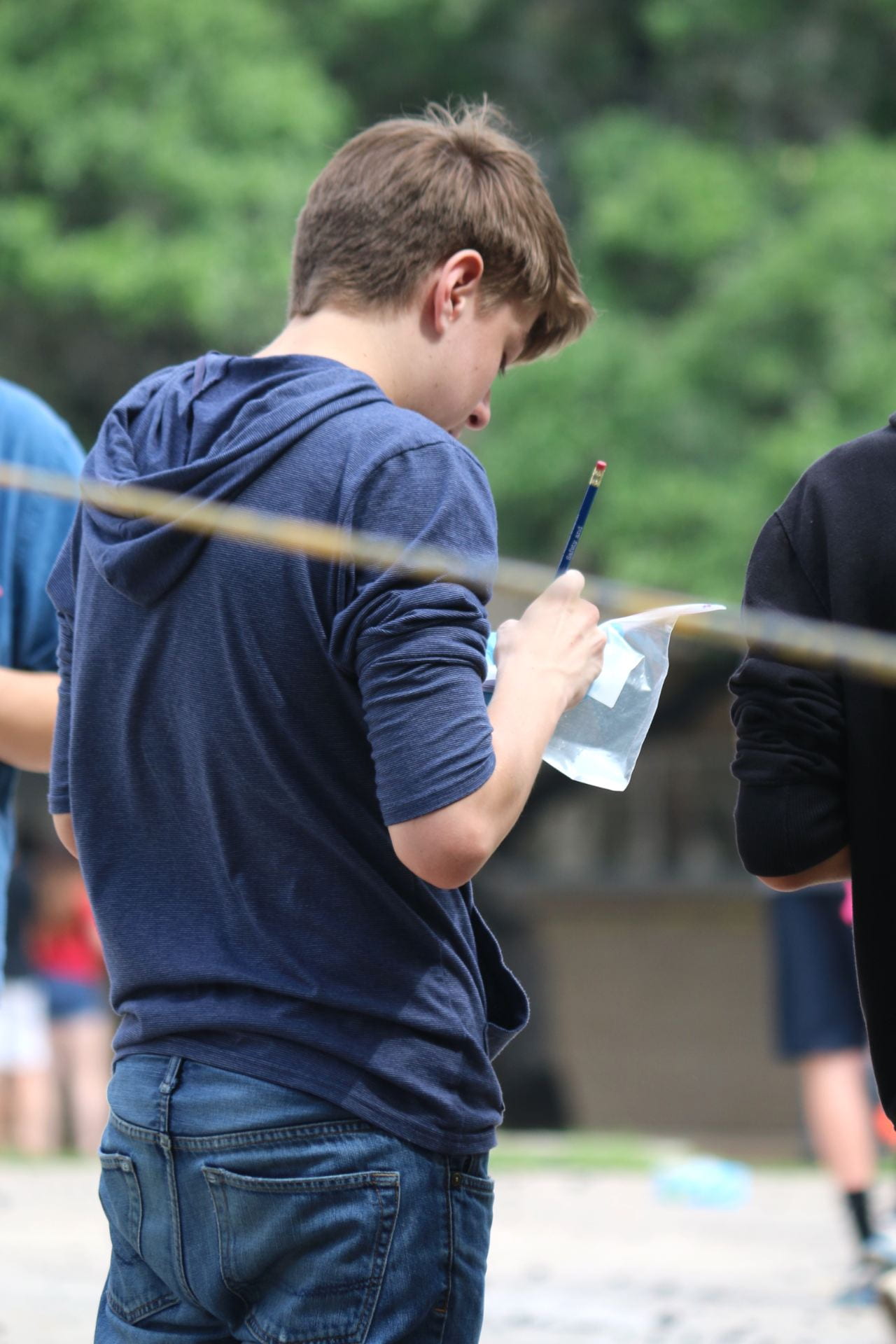 A young person wearing a navy hoodie is standing outdoors, focused on writing on a piece of paper while holding a pen, with blurred figures in the background.