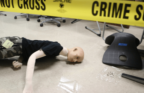 A crime scene setup featuring a mannequin lying on the floor near a chair, with yellow police tape reading 'DO NOT CROSS' in the foreground.