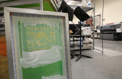 A screen printing setup featuring a partially coated screen in the foreground, with various printing equipment and tools visible in the background.