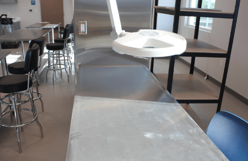 A modern kitchen space featuring a stainless steel countertop, a white overhead light fixture, and bar stools in the background.