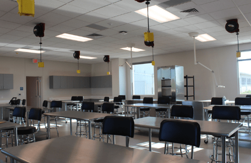 A spacious classroom featuring multiple tables and chairs, with overhead equipment and large windows allowing natural light to enter.