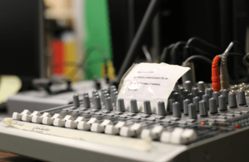 A close-up view of a sound mixing console with various knobs and a note placed on it, set against a blurred background of equipment.