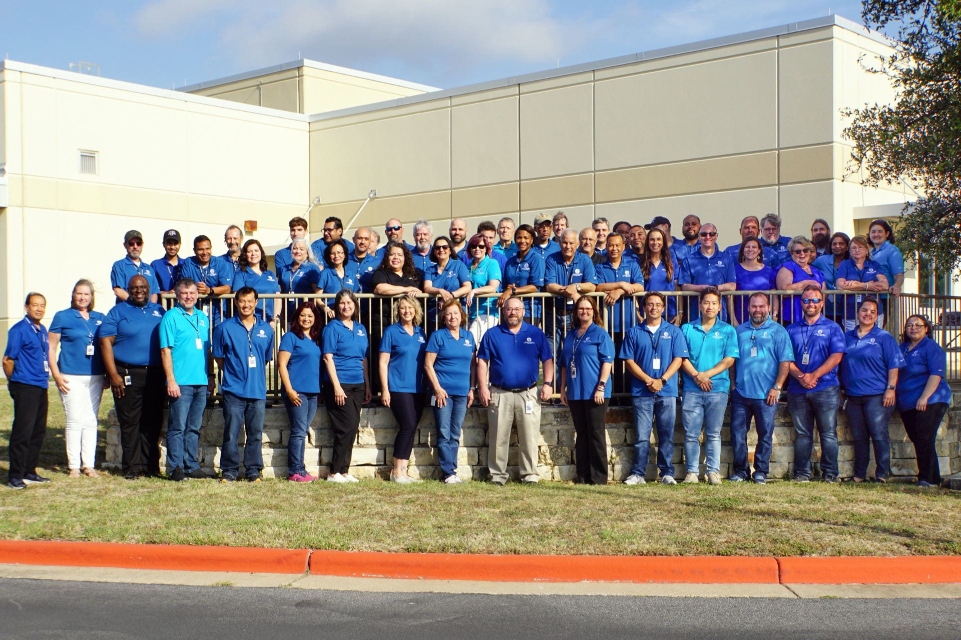 A large group of people wearing blue shirts stands together on steps outside a building, with some individuals on a higher level and others on the ground, all smiling for the camera.