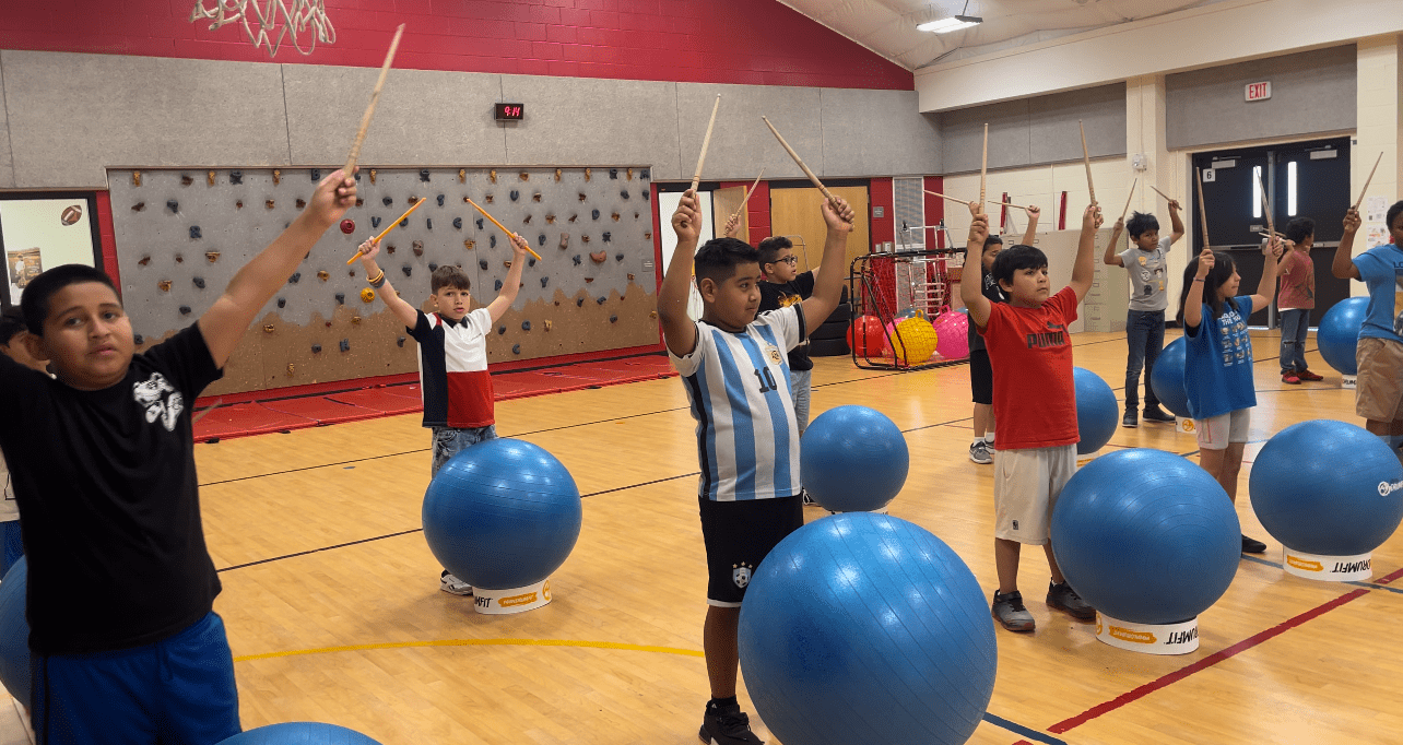 A group of children in a gymnasium are standing on blue exercise balls while holding drumsticks in the air, participating in a rhythmic activity.