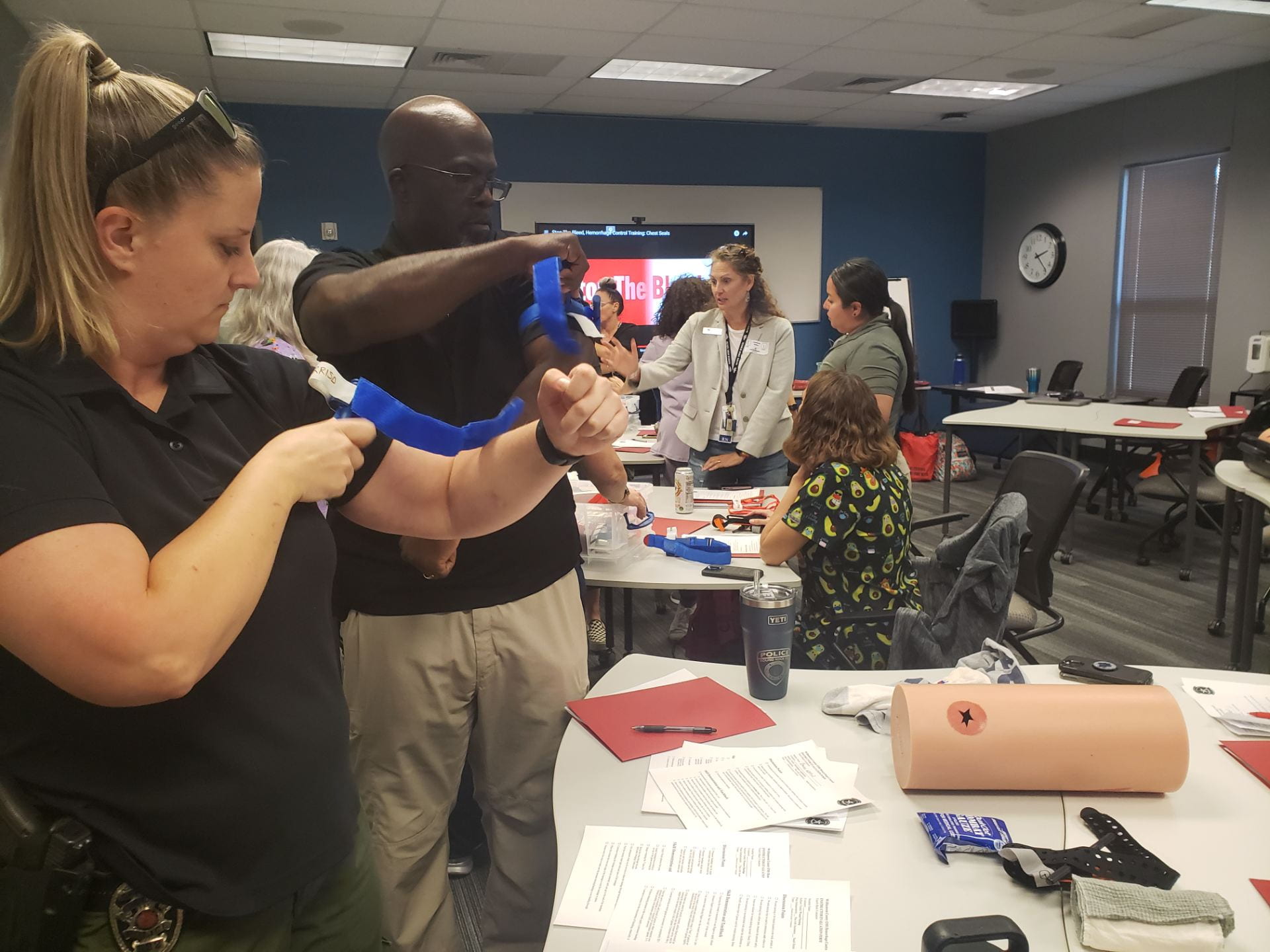 A group of people participating in a training session, with one woman applying a tourniquet on a man's arm while others observe and engage in the background.