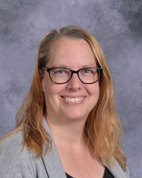 A woman wearing glasses and a gray cardigan smiles warmly, exuding a friendly and approachable demeanor.