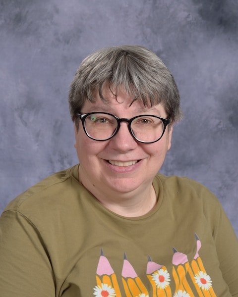 A woman wearing glasses and a green shirt smiles warmly, exuding a friendly and approachable demeanor.