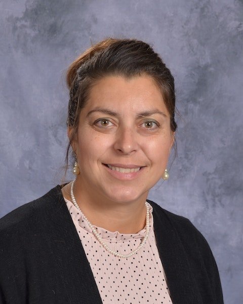 A woman wearing a pink shirt and a black cardigan, standing confidently with a neutral background.