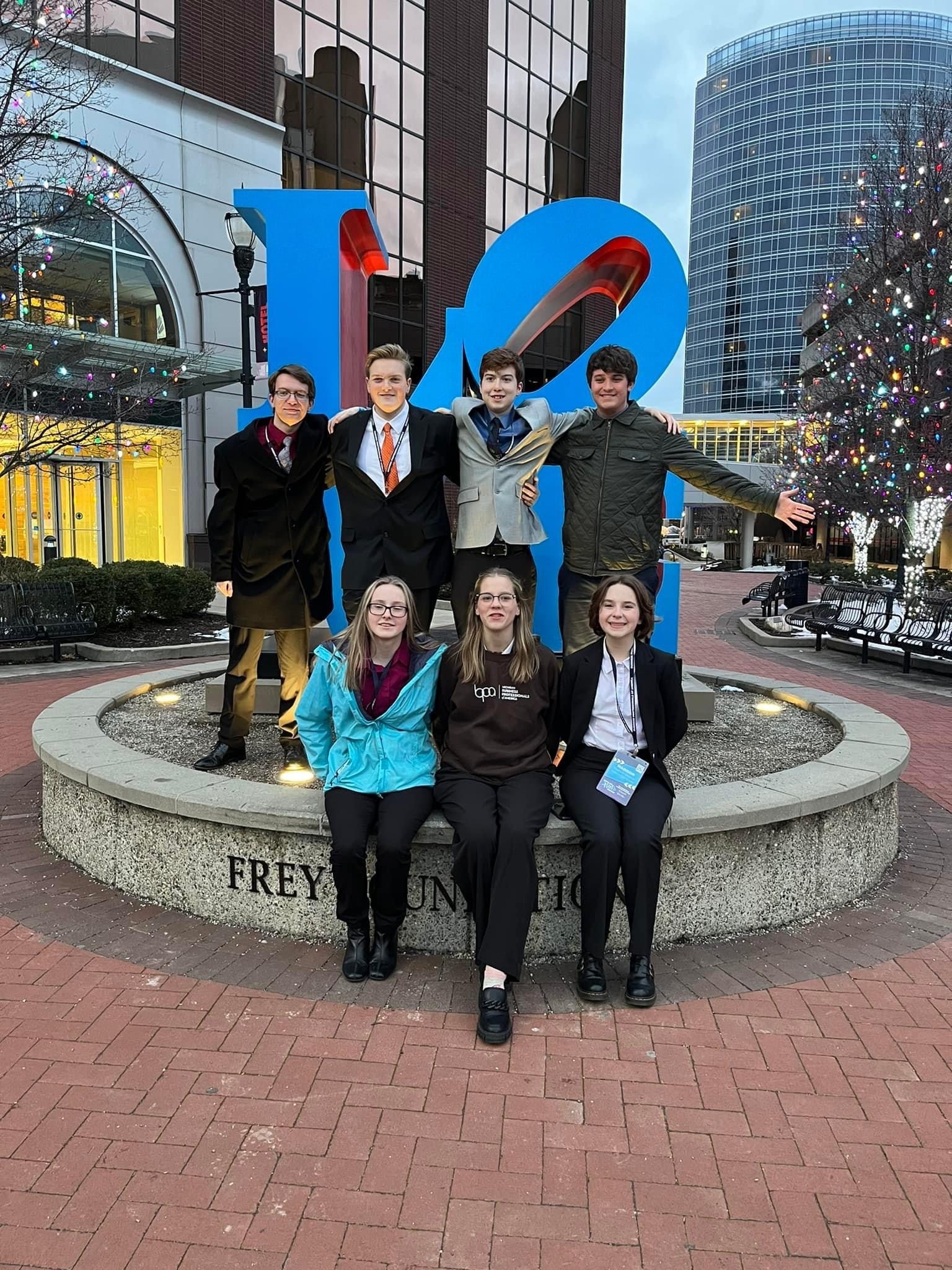 A group of seven people poses in front of a large, colorful sculpture in an urban setting, with buildings and festive lights in the background.