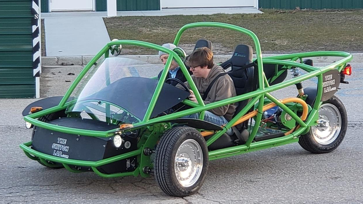 A person sits in a unique, open-frame green vehicle with a transparent windshield and two seats, showcasing its unconventional design.