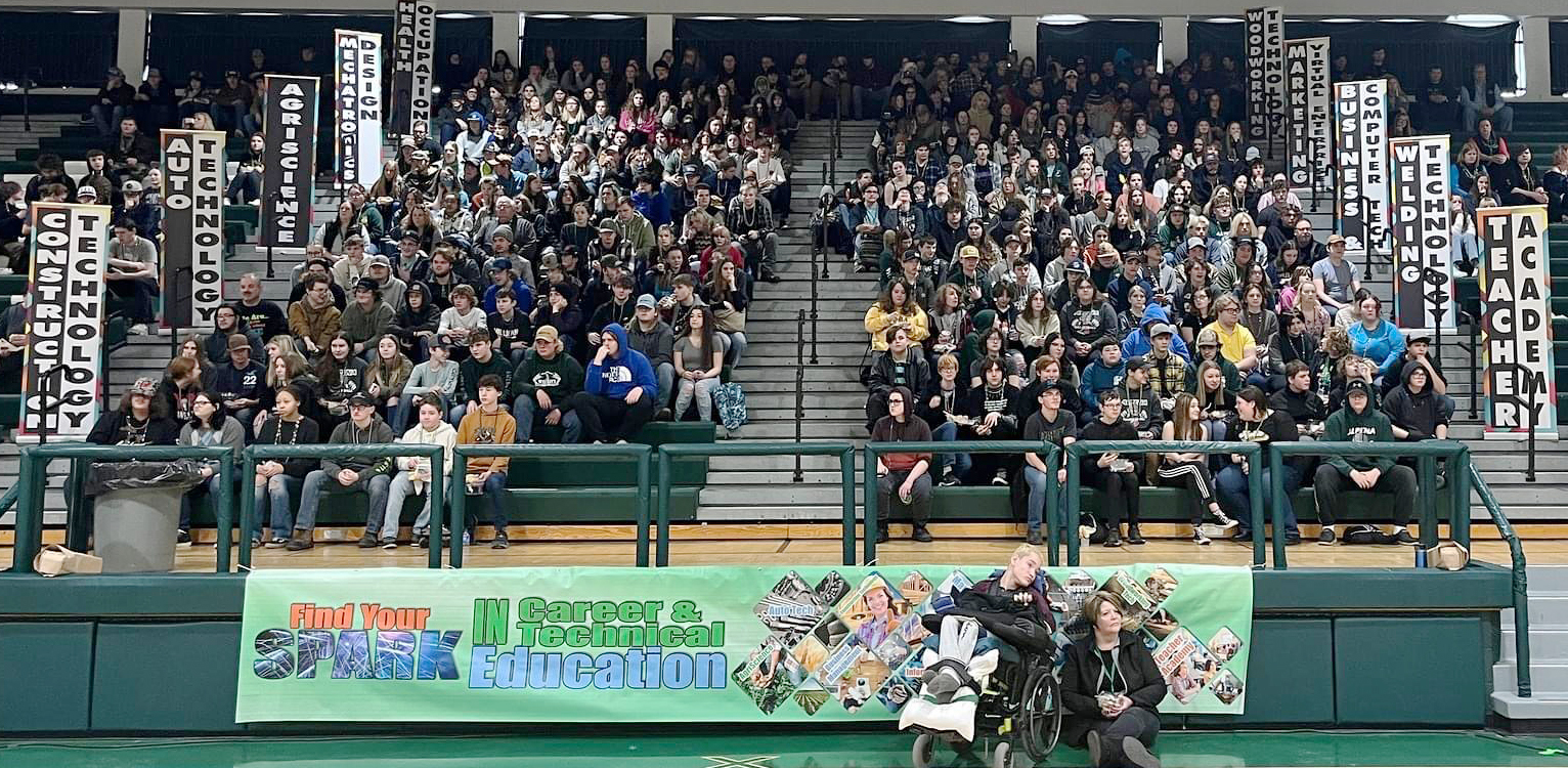 A large crowd of people fills a gymnasium, with banners and signs visible in the background, indicating a spirited event or competition.