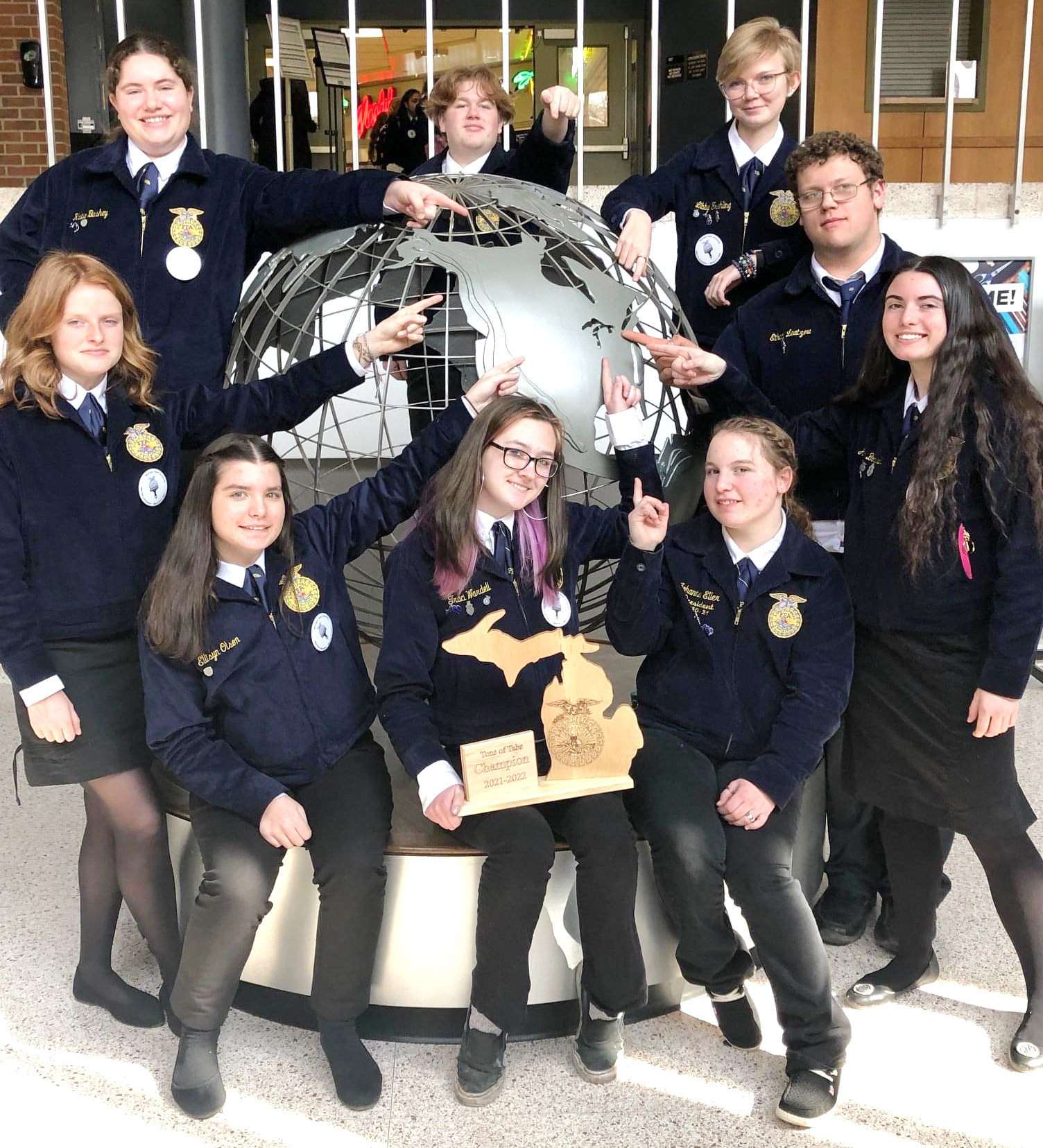 A group of nine students in blue jackets poses around a globe sculpture, with some pointing at it and others holding a wooden plaque, all smiling in a school setting.