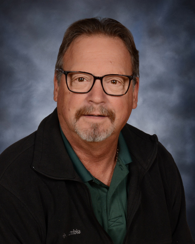 A middle-aged man with glasses and a beard is smiling at the camera, wearing a black jacket over a green shirt, set against a softly blurred blue background.
