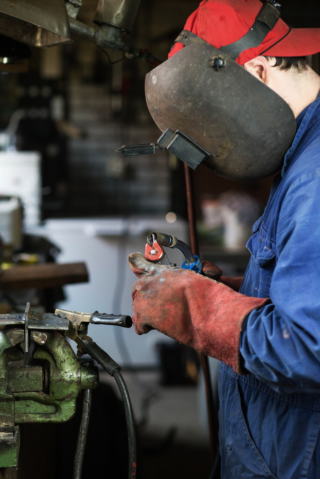 A person wearing protective gear and a welding mask is working on a metal piece in a workshop, with tools and equipment visible in the background.