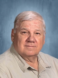 A senior man with short, light-colored hair and a slight smile, wearing a light-colored collared shirt, against a blue background.